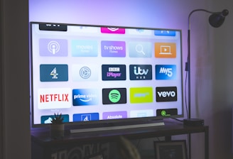 A flat-screen television is displaying a variety of streaming app icons, including Netflix, Prime Video, Spotify, and ITV Hub among others. The TV is placed on a media console with a small potted plant beside it. A tall lamp stands to the right, and a faint, ambient light casts a purple hue on the wall above the TV.