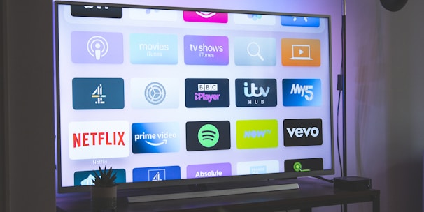 A flat-screen television is displaying a variety of streaming app icons, including Netflix, Prime Video, Spotify, and ITV Hub among others. The TV is placed on a media console with a small potted plant beside it. A tall lamp stands to the right, and a faint, ambient light casts a purple hue on the wall above the TV.