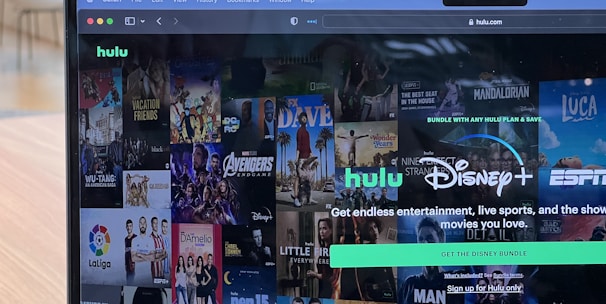 A laptop screen displays a web page featuring a mix of movie and TV show covers, highlighting popular titles and a Hulu and Disney+ bundle promotion. There are visible icons and images of films and shows alongside promotional text offering entertainment and live sports.